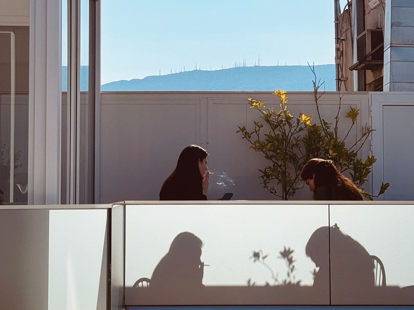 Two women at a rooftop breakfast table in Athens, their shadows cast on the frosted glass railing, a hill lined with antennas beyond. Photograph by Christopher Kulawik.