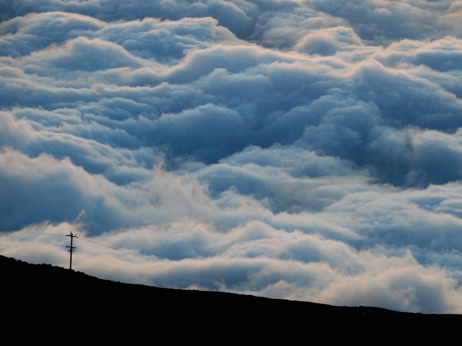 A sea of clouds below the slopes of Haleakalā, with a lone utility pole on the dark ridgeline. Photograph by Christopher Kulawik.