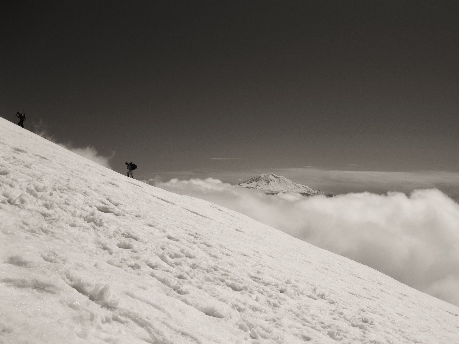 Two climbers on the snowfield of Mount St. Helens above a sea of cloud, a distant snow-capped peak on the horizon. Photograph by Christopher Kulawik.
