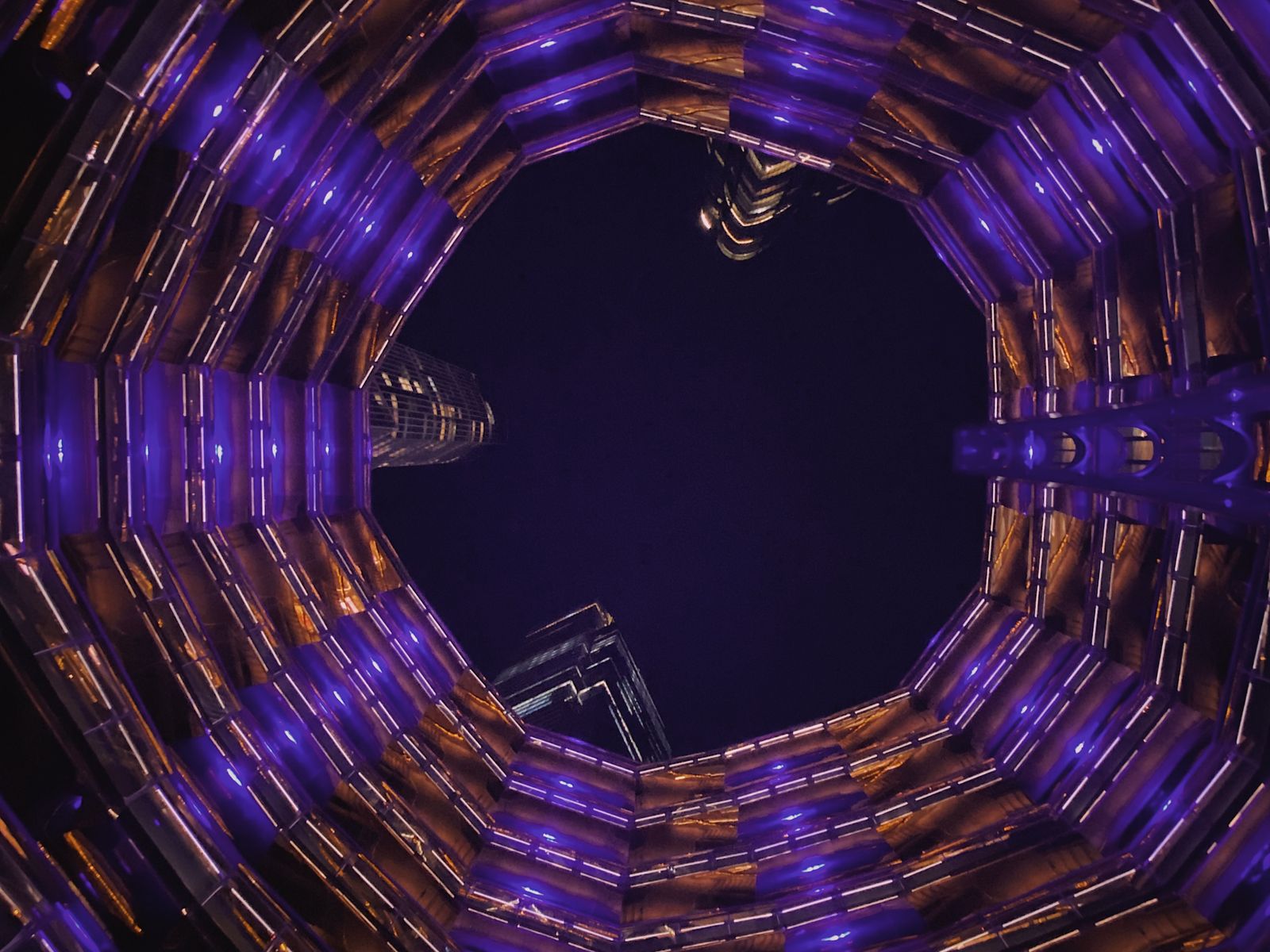 Looking straight up through the honeycomb of the Vessel at Hudson Yards at night, lit purple and copper, skyscrapers framed in the opening. Photograph by Christopher Kulawik.